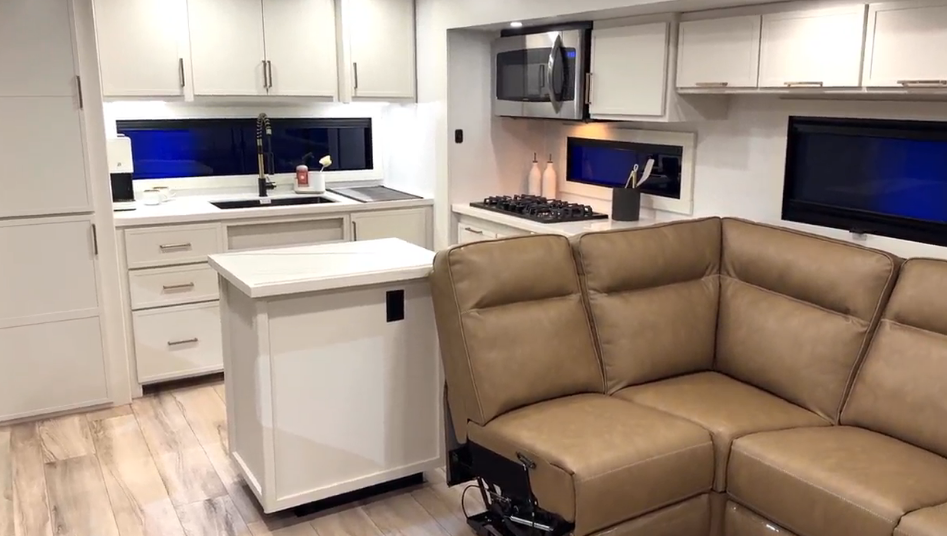 Interior view of the Brinkley Model I 280 showcasing the open-concept rear kitchen and living area with opposing slides. The image features a solid-surface island, tan leather L-shaped recliner seating, stainless steel convection microwave, and three-burner cooktop—highlighting the RV’s residential design, warm lighting, and premium finishes.