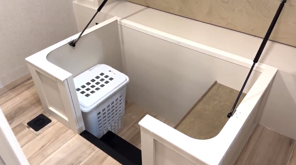 Under-bed storage compartment in the Brinkley Model I 280 featuring a deep step-down design with built-in clothing rack supports and a laundry basket. Gas struts hold the bed frame open, revealing ample organized space ideal for clothing, linens, or pet supplies—showcasing Brinkley’s innovative use of every inch.