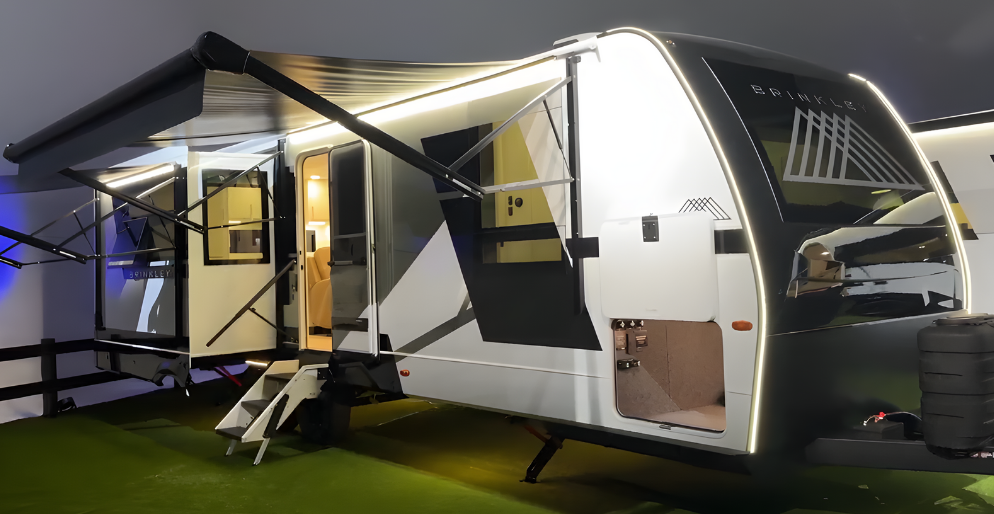 Exterior view of the Brinkley Model I 280 travel trailer showcasing its modern automotive styling with a ¾ front cap and illuminated Brinkley logo. The dual awnings, large slide-outs, and frameless windows highlight its luxury design, while the wide entry steps and open storage bay emphasize accessibility and premium construction.