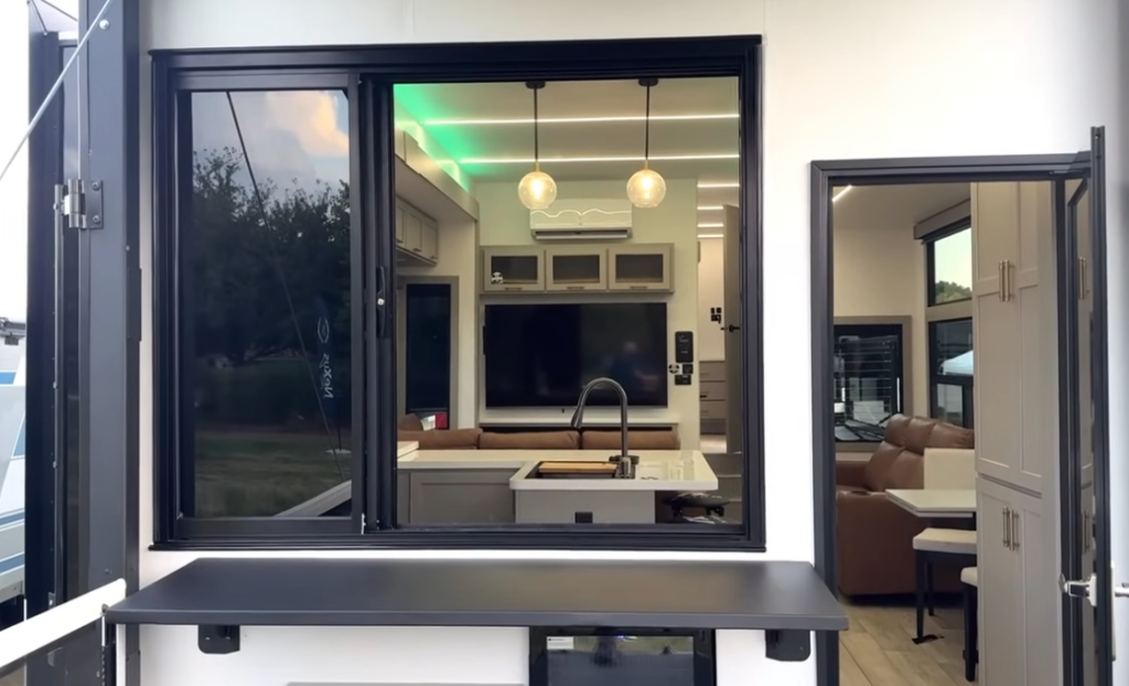 Outdoor view into the 2026 Axiom Imperium I3800 fifth wheel kitchen showing large sliding glass windows, island sink, pendant lights, modern cabinetry, and seamless access between the rear patio and interior living area.