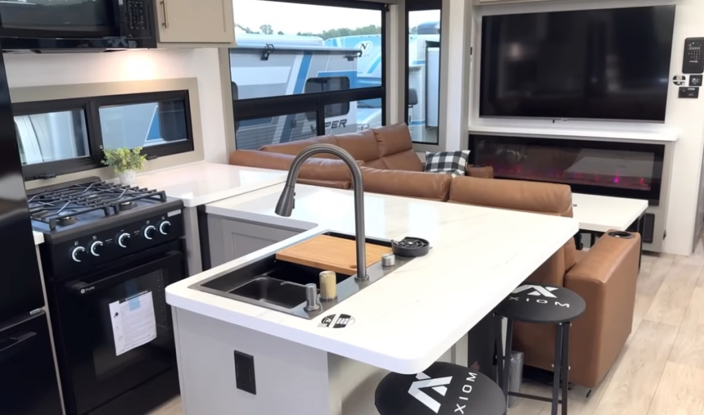 Spacious kitchen and living area inside the 2026 Axiom Imperium I3800 fifth wheel featuring a center island with farmhouse sink, gas range, GE refrigerator, leather sectional, floating dining table, large TV, and modern electric fireplace.