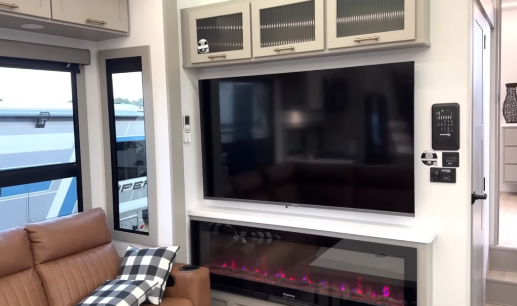 Entertainment center inside the 2026 Axiom Imperium I3800 featuring a large flat-screen TV, modern electric fireplace with color lighting, upper storage cabinets, and a leather sofa with plaid pillow in a luxury RV living space.