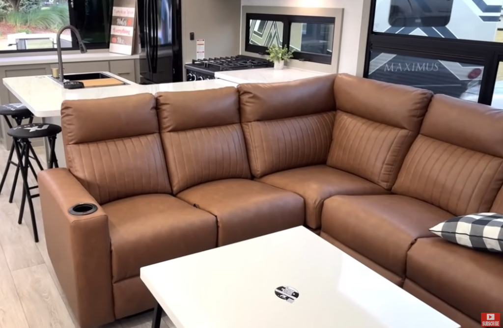Luxury RV interior featuring a tan leather sectional sofa with built-in cupholder, modern white table, and contemporary kitchen in the background with island sink, barstools, and stainless steel appliances inside the 2026 Axiom Imperium I3800 fifth wheel.