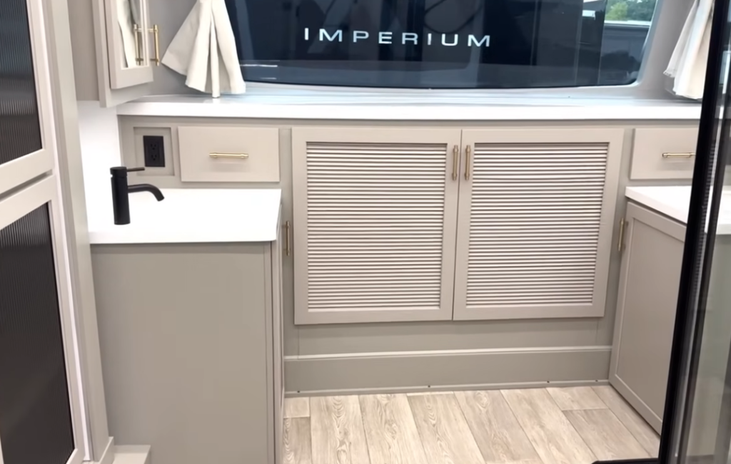 Bedroom vanity area inside the 2026 Axiom Imperium I3800 fifth wheel featuring soft-close cabinetry with louvered doors, black faucet, gold hardware, and large rear window with “Imperium” branding visible outside.