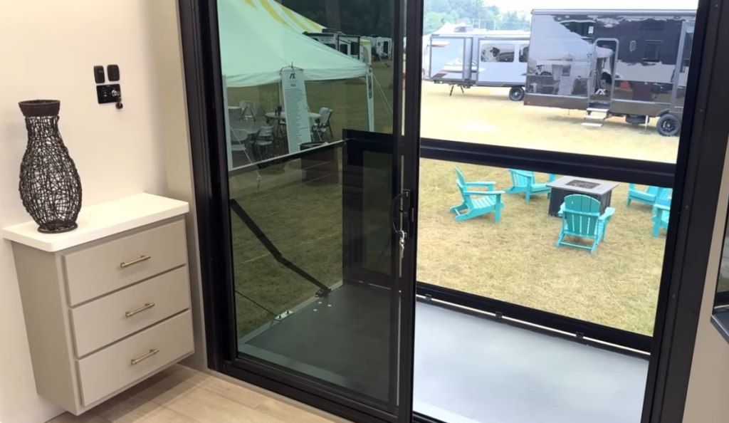Bedroom balcony of the 2026 Axiom Imperium I3800 fifth wheel with sliding glass doors opening to an elevated outdoor deck, featuring glass railings and a view of turquoise patio chairs around a fire pit.