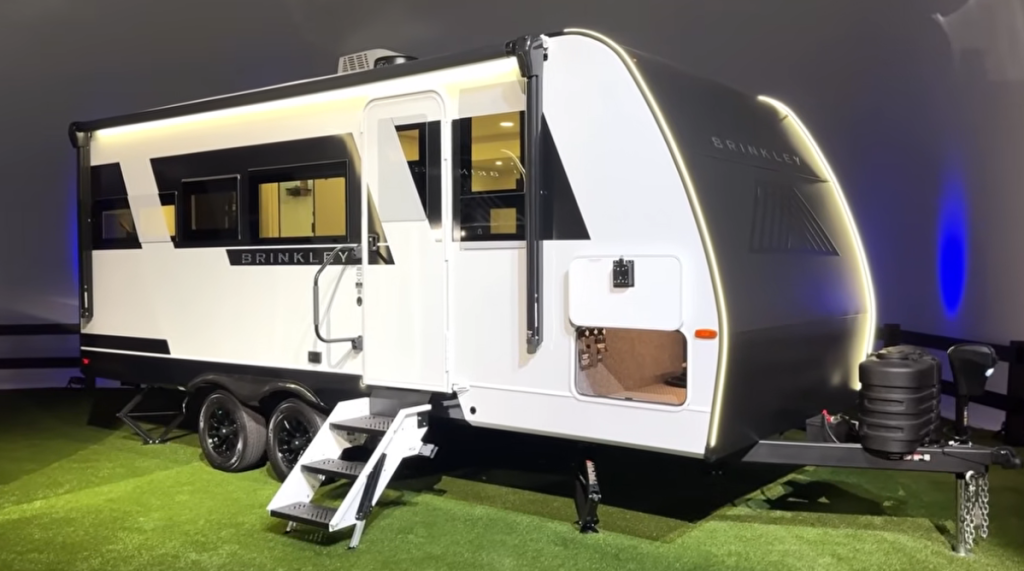 Brinkley Model Ix 20X travel trailer on display with painted aluminum exterior, dual-axle frame, LED fusion lighting, and solid entry steps, highlighting the compact half-ton towable design.