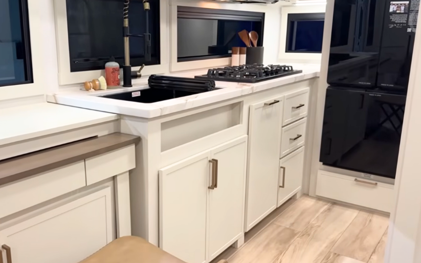 Brinkley Model Ix 20X kitchen interior with marble-look countertops, stainless farmhouse sink, three-burner cooktop, modern cabinetry, and 12V refrigerator inside the compact luxury travel trailer.