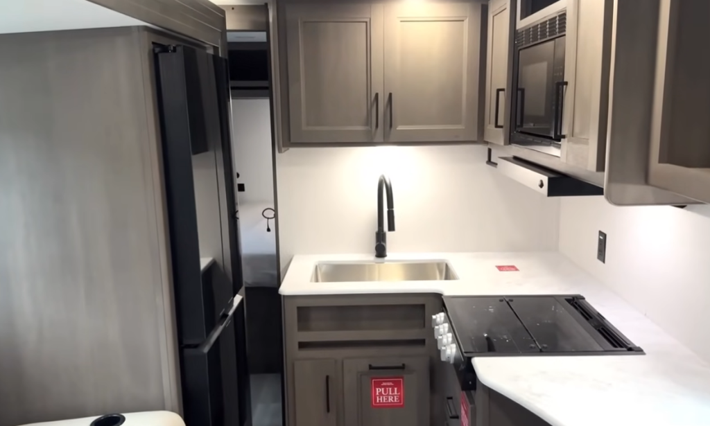 Grand Design Transcend Xplor 22RBX kitchen with stainless steel sink, modern cabinetry, microwave, three-burner stove, and residential refrigerator.