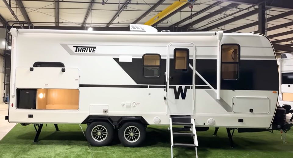 Winnebago Thrive travel trailer side exterior view showing modern black and white design, dual-axle frame, entry steps, large awning, and outdoor storage compartment with built-in mini fridge—designed for style, comfort, and functionality.