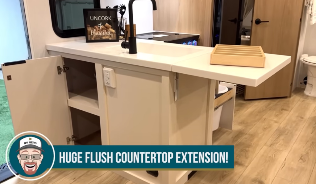 Winnebago Thrive RV kitchen island with massive flush countertop extension, matte black faucet, white cabinetry with open storage, and wood-look flooring, highlighting the trailer’s smart space design and functional meal prep area.