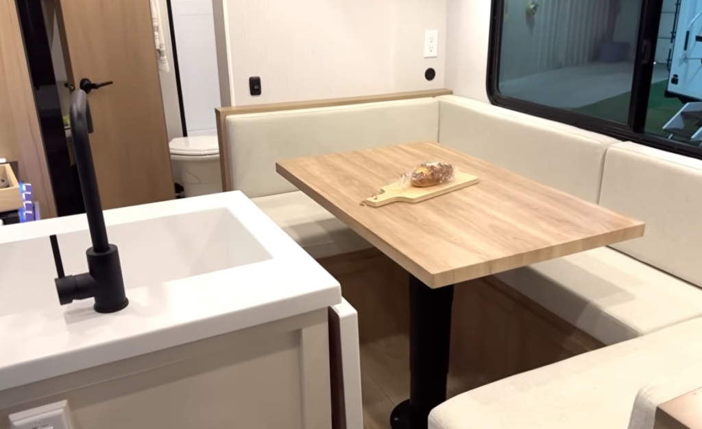 Winnebago Thrive travel trailer interior featuring a modern U-shaped dinette with light beige cushions, natural wood table, and white farmhouse-style kitchen sink with matte black faucet, showcasing open-concept RV dining and kitchen design.