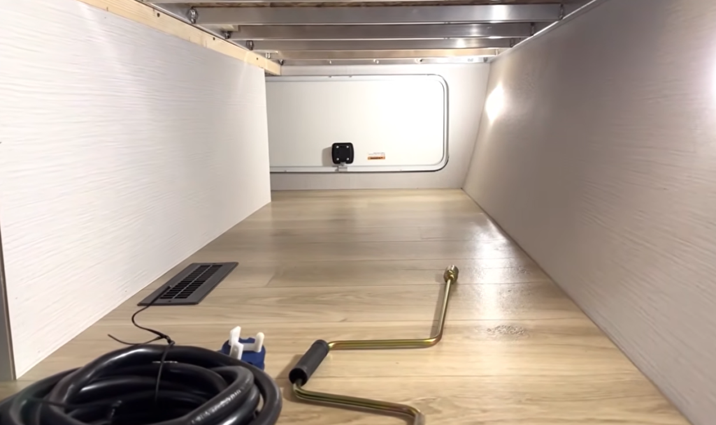 Pass-through storage compartment inside a Winnebago Thrive travel trailer, showcasing clean laminated walls, LED lighting, and organized utility connections with hoses and tools for easy access and setup.