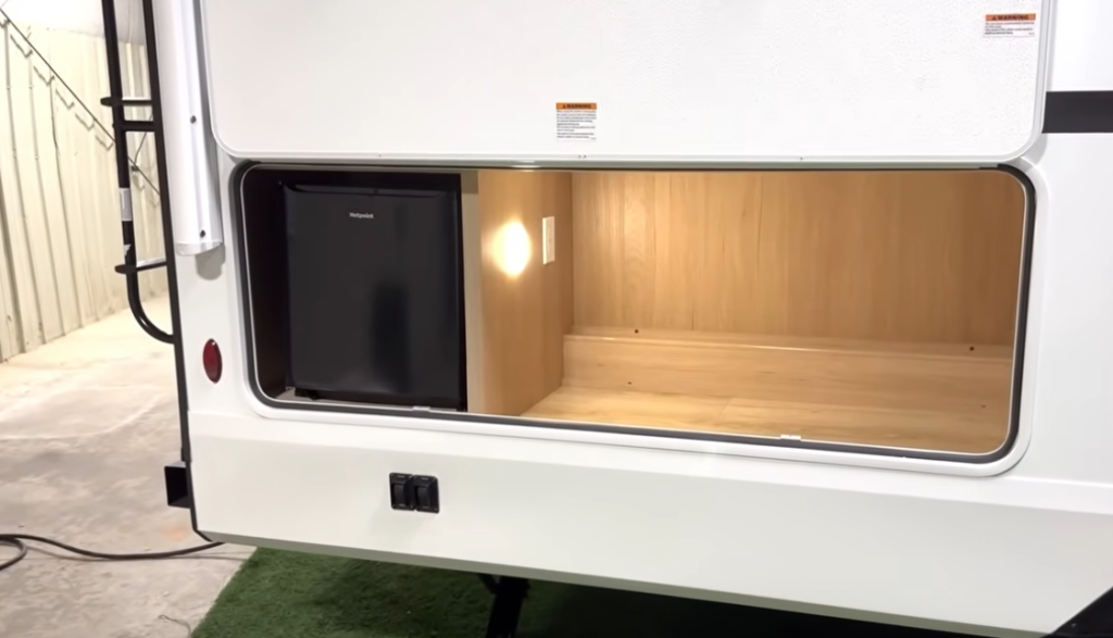 Exterior storage compartment of a Winnebago Thrive travel trailer, featuring a built-in Hotpoint mini fridge and spacious lighted cargo area with wood-grain finish for convenient outdoor access and organization.