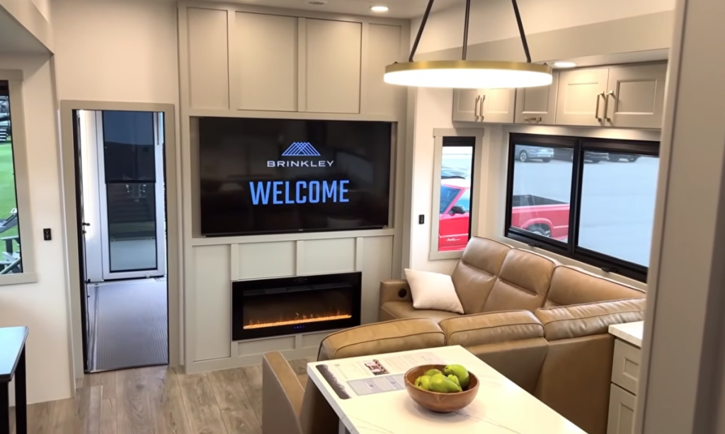 Brinkley Model G Series luxury RV living area featuring a tan leather sectional sofa, large LED TV with ‘Brinkley Welcome’ display, modern electric fireplace, pendant lighting, and panoramic windows for a bright residential feel.