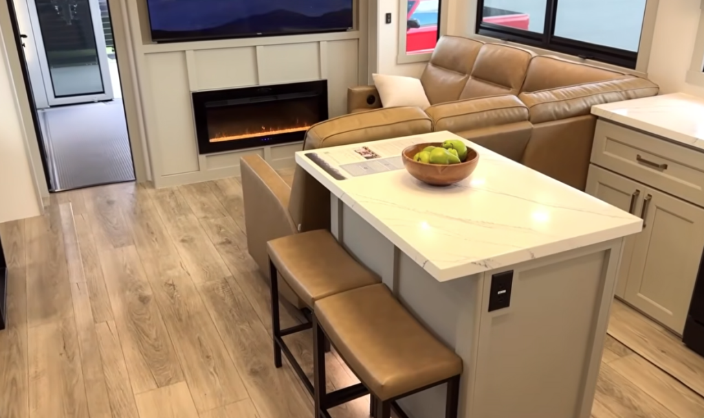“Brinkley Model G Series RV interior featuring a modern island dining bar with leather stools, quartz-style countertop, built-in fireplace, and tan sectional sofa, blending luxury design with functional living space.