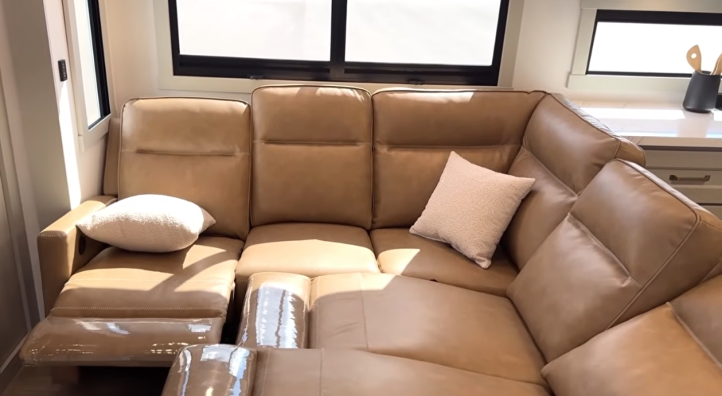 Brinkley Model G Series RV living room showcasing a plush tan leather sectional with dual recliners, accent pillows, and large panoramic windows for a bright, upscale residential lounging experience.
