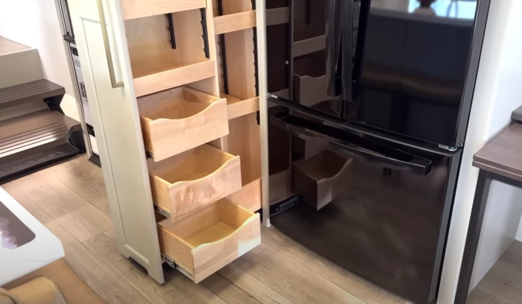 Brinkley Model G Series RV kitchen storage with dual pull-out pantries beside a black residential refrigerator, featuring adjustable shelves and deep wood drawers for efficient food and cookware organization.