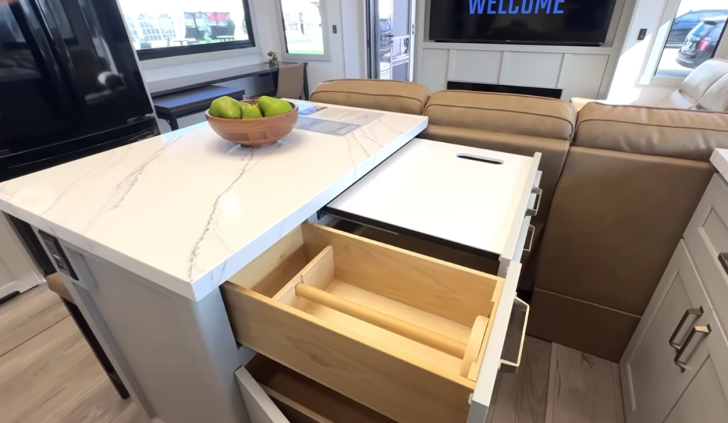 Brinkley Model G Series RV kitchen island featuring a marble-look countertop, built-in cutting board, deep storage drawers with utensil organizer, and modern open-concept layout facing the living area with fireplace and sectional.