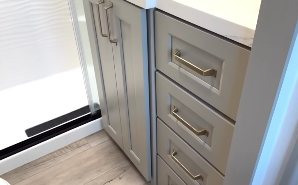 Brinkley Model G Series RV bathroom cabinetry with soft-close drawers, gold hardware, and solid-surface countertop beside a walk-in shower, combining luxury design with practical storage.
