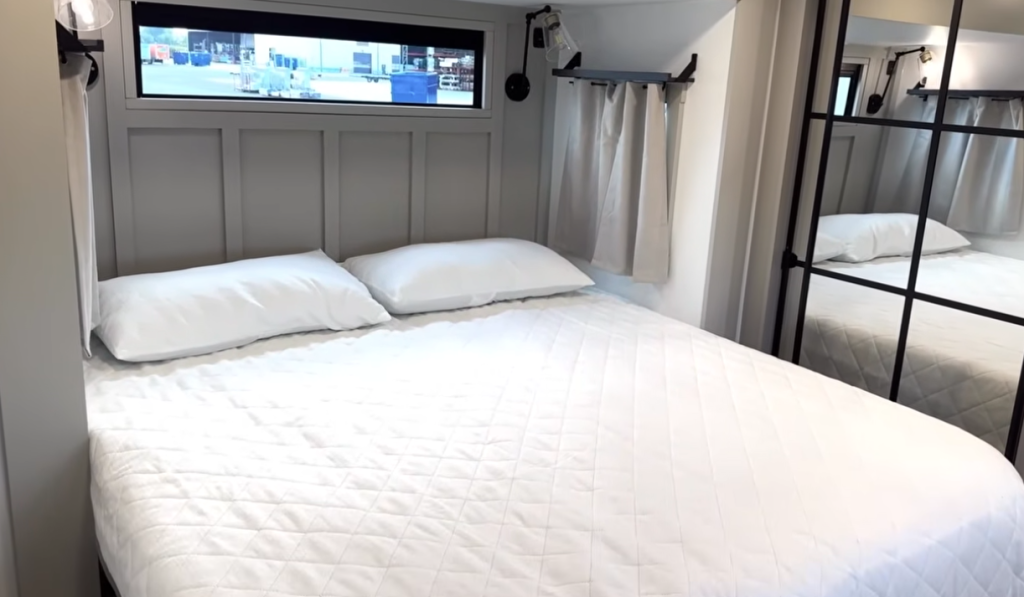 Brinkley Model G Series RV bedroom featuring a residential king bed with quilted bedding, modern reading sconces, high transom window, and full-length mirrored wardrobe for a bright, spacious retreat.