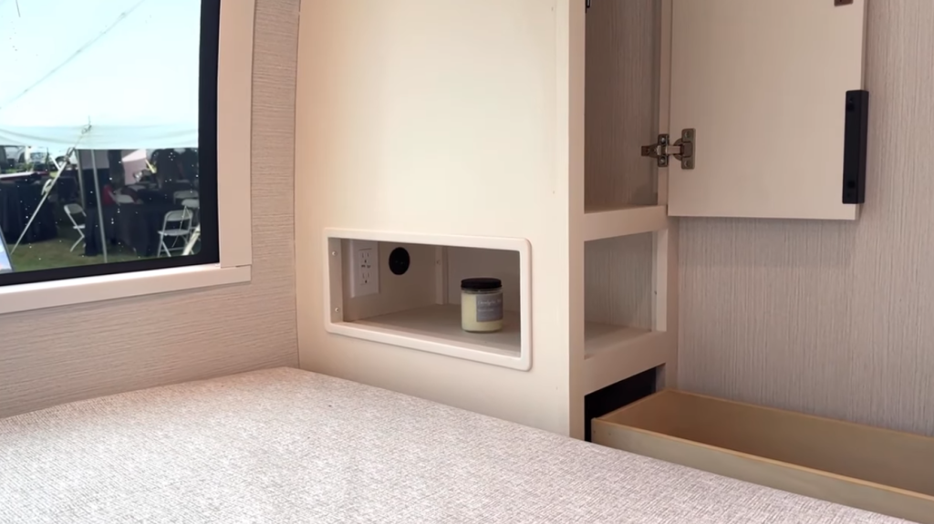 Close-up of the 2026 Winnebago Thrive 22MLS bedroom storage with built-in nightstand shelf, electrical outlet, cabinet, and drawer beside the Olympic queen bed for convenient organization.