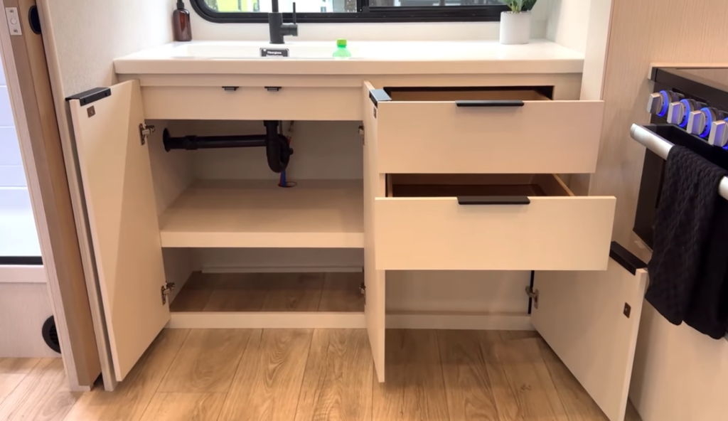 2026 Winnebago Thrive 22MLS kitchen storage with open cabinets and drawers under the sink, showing ample space for cookware and essentials in a modern, light-toned design.