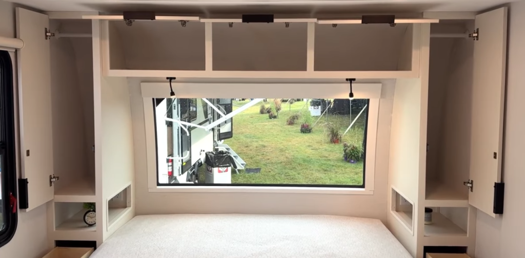 2026 Winnebago Thrive 22MLS bedroom with large panoramic front window, overhead storage cabinets, side wardrobes, and integrated lighting for a bright, modern sleeping space.