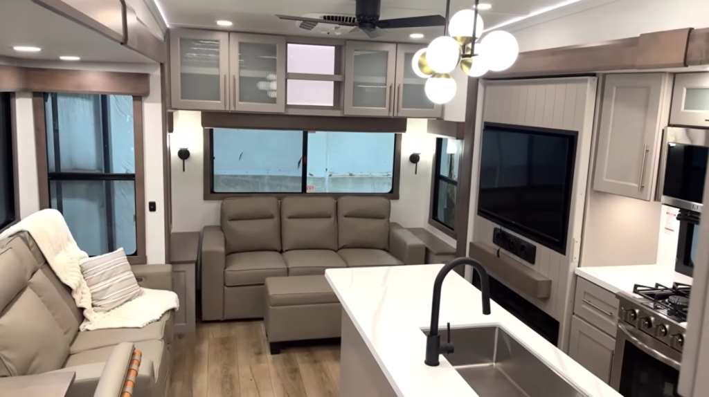 “Alliance Paradigm fifth wheel RV interior featuring dual recliners, a sofa, entertainment center with TV and fireplace, modern kitchen island with black faucet, and pendant lighting for a bright residential feel.