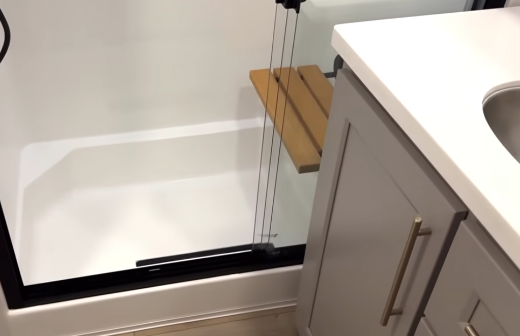 Alliance Paradigm fifth wheel bathroom with modern vanity, solid-surface countertop, and walk-in shower featuring a fold-down teak bench for comfort and accessibility.