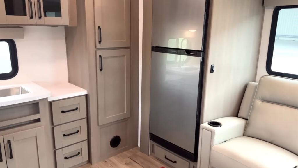 2026 Grand Design Reflection 150 270BN kitchen featuring a double-door refrigerator, pantry storage, and modern cabinetry beside the living area recliners.