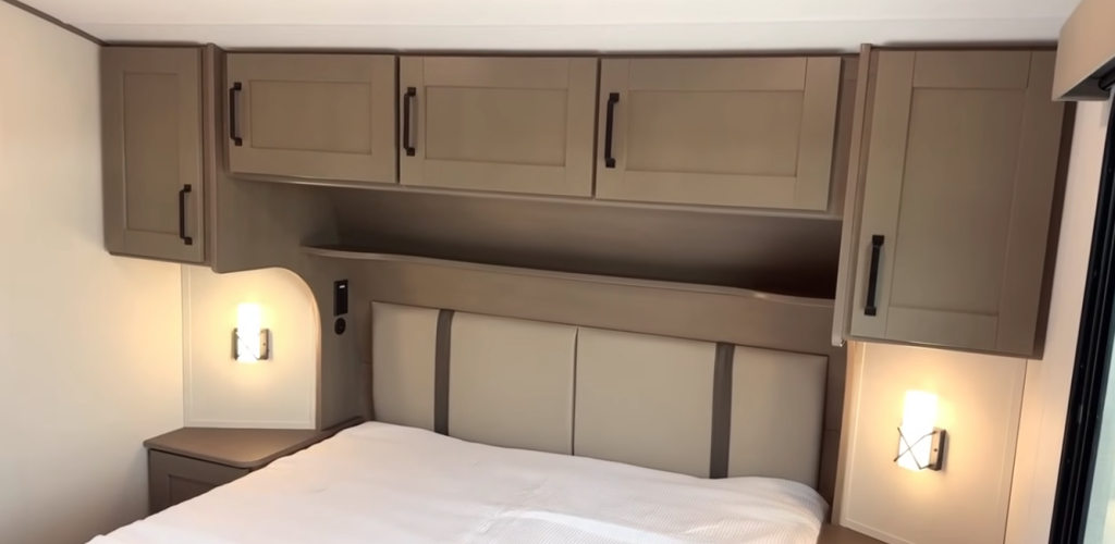 2026 Grand Design Reflection 150 270BN bedroom with overhead cabinetry, bedside storage, and soft reading lights above a padded headboard for a modern, cozy retreat.