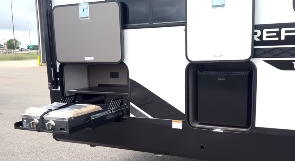 2026 Grand Design Reflection 150 270BN exterior camp kitchen with pull-out dual-burner stove, mini refrigerator, and storage compartments for convenient outdoor cooking.