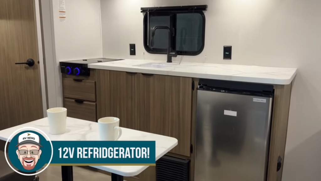 Forest River Wildwood FSX compact RV kitchen with Everchill 12V refrigerator, two-burner cooktop, stainless sink, and modern wood cabinetry for efficient small-space cooking.