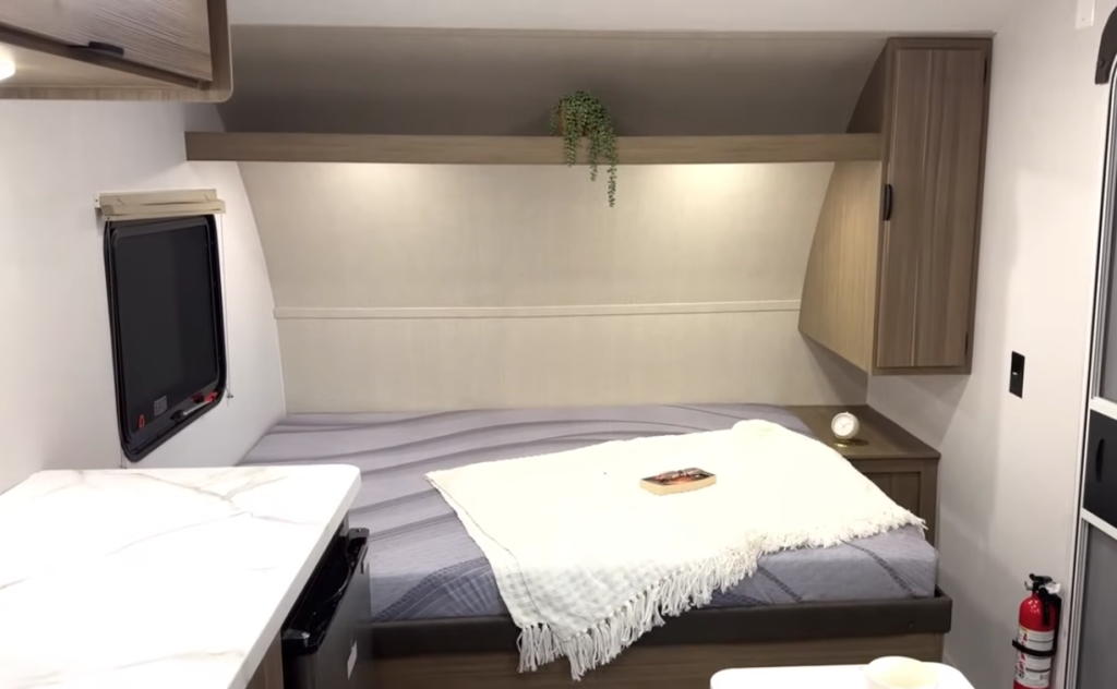 Forest River Wildwood FSX RV bedroom with corner cabinetry, nightstand, side window, and cozy full-size bed topped with a throw blanket and book.