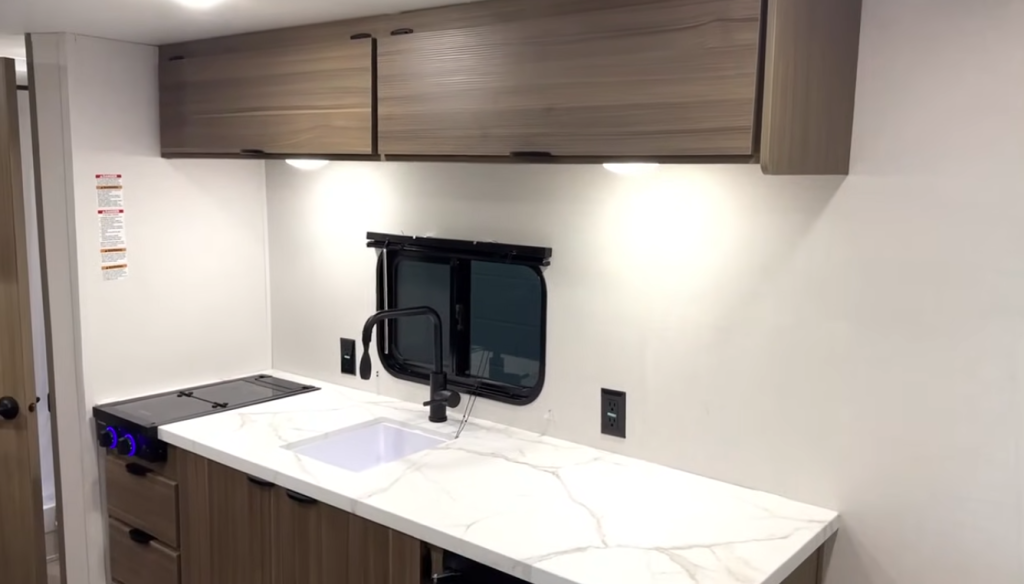 Forest River Wildwood FSX RV kitchen interior featuring marble-look countertop, single-basin sink with black faucet, two-burner cooktop, and modern overhead storage cabinets.