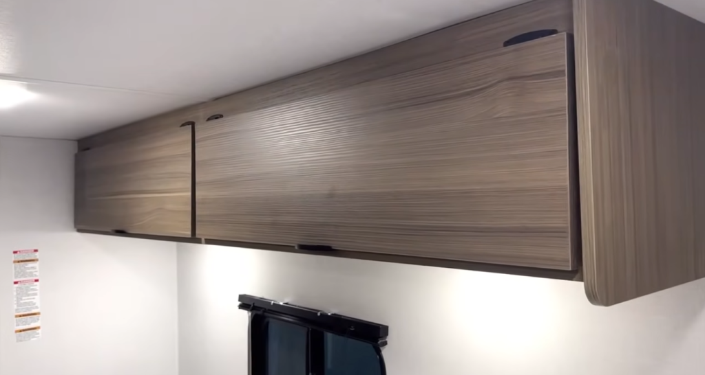 Overhead wood-grain storage cabinets inside the Forest River Wildwood FSX travel trailer, providing modern, lightweight storage above the kitchen window.
