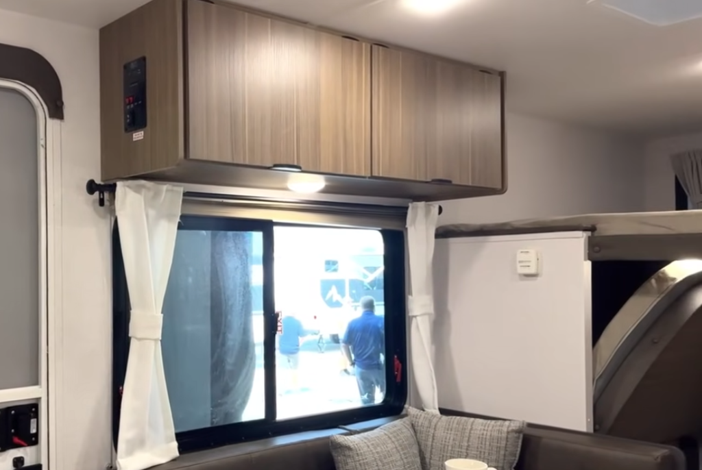 Forest River Wildwood FSX RV interior showing upper storage cabinets, large window with white curtains, and dinette area beside the convertible Murphy bed setup.
