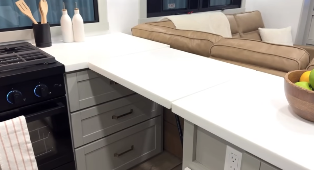 Brinkley Model Z fifth wheel kitchen with white solid-surface countertops, an extended counter workspace, built-in oven, and modern cabinetry beside a tan leather sectional for a cohesive open-concept design.