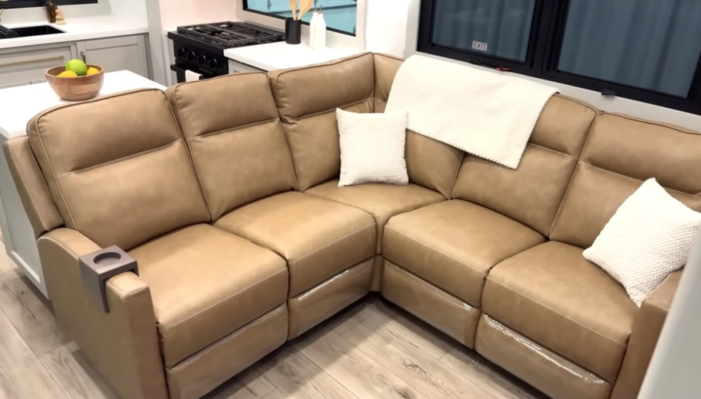 Brinkley Model Z fifth wheel interior showing a large tan leather L-shaped sofa with built-in cupholder, soft accent pillows, and modern RV kitchen in the background for a luxury residential feel.