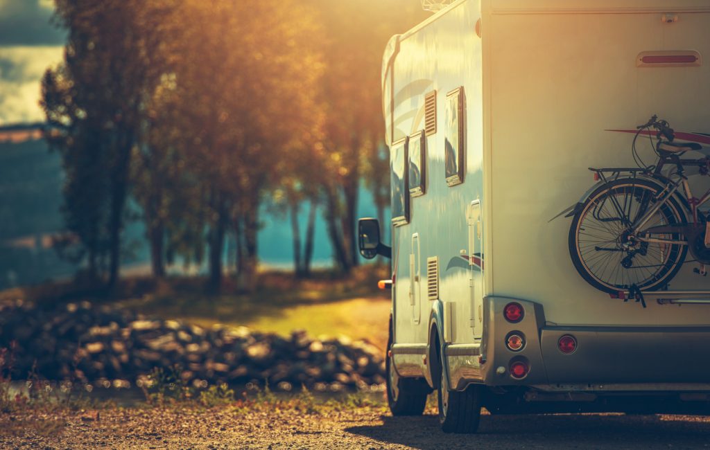Rear view of a motorhome parked by a scenic lakeside at sunset with bicycles mounted on the back, surrounded by trees and golden light reflecting off the water.