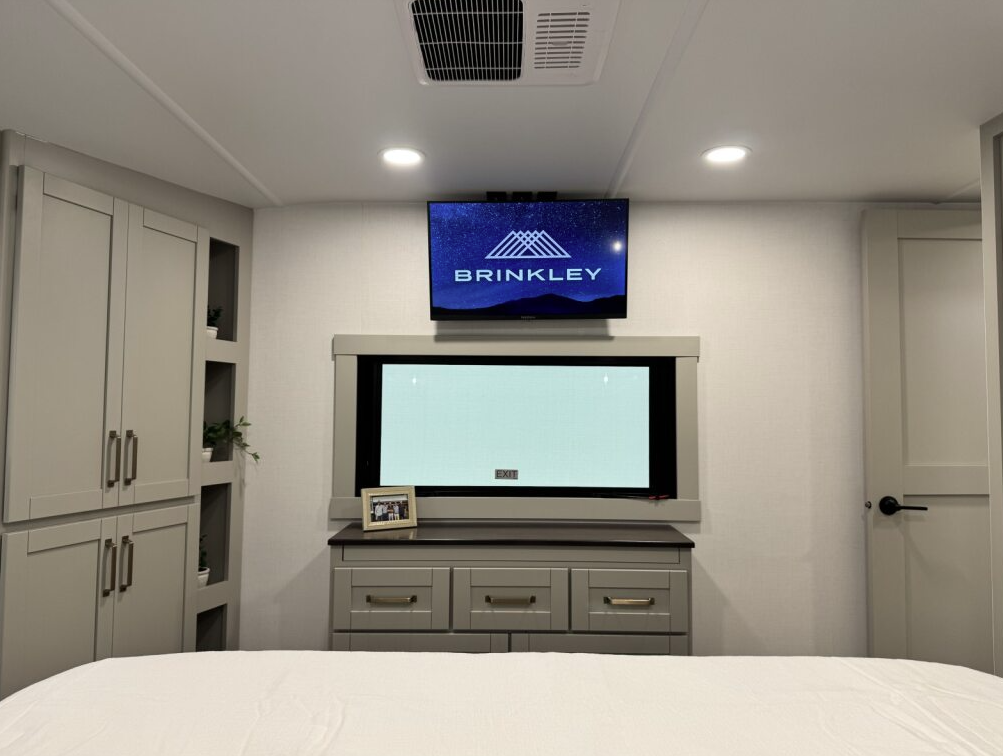 Brinkley Model Z 3600 bedroom featuring a mounted TV above a large window, dresser drawers with gold handles, and built-in cabinetry with open shelving and modern lighting.