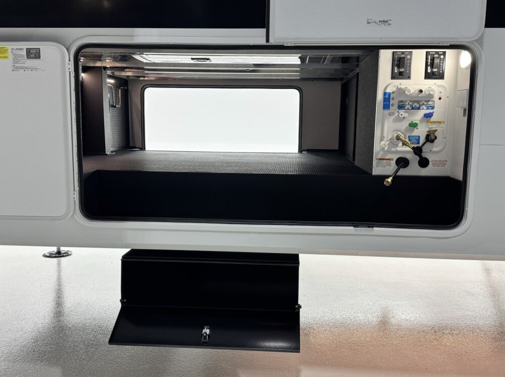 Pass-through storage compartment on a Brinkley fifth wheel RV featuring a clean utility panel with labeled water and power connections, bright LED lighting, and durable flooring for organized storage.