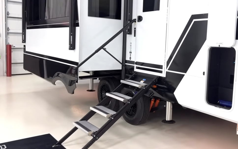 Exterior view of a Brinkley fifth wheel RV entry featuring sturdy quad-step stairs, large entry door, and pass-through storage compartment for easy access and setup.
