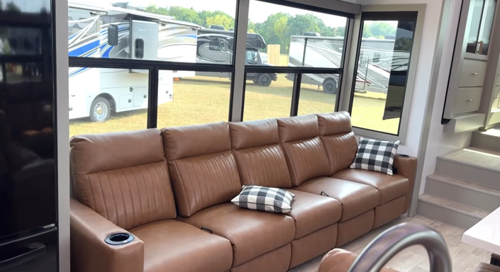 Interior of the 2026 Axiom Vendetta 4250 fifth wheel featuring a large tan leather reclining sofa with plaid accent pillows, panoramic windows, and modern cabinetry for a bright, spacious living area.