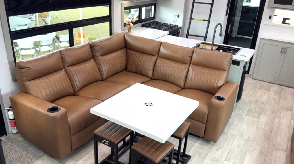 Modern living area inside the 2026 Axiom Vendetta 4250 fifth wheel toy hauler, featuring an L-shaped tan leather recliner sofa, removable dining table with matching stools, and a full kitchen with island sink and stainless appliances.
