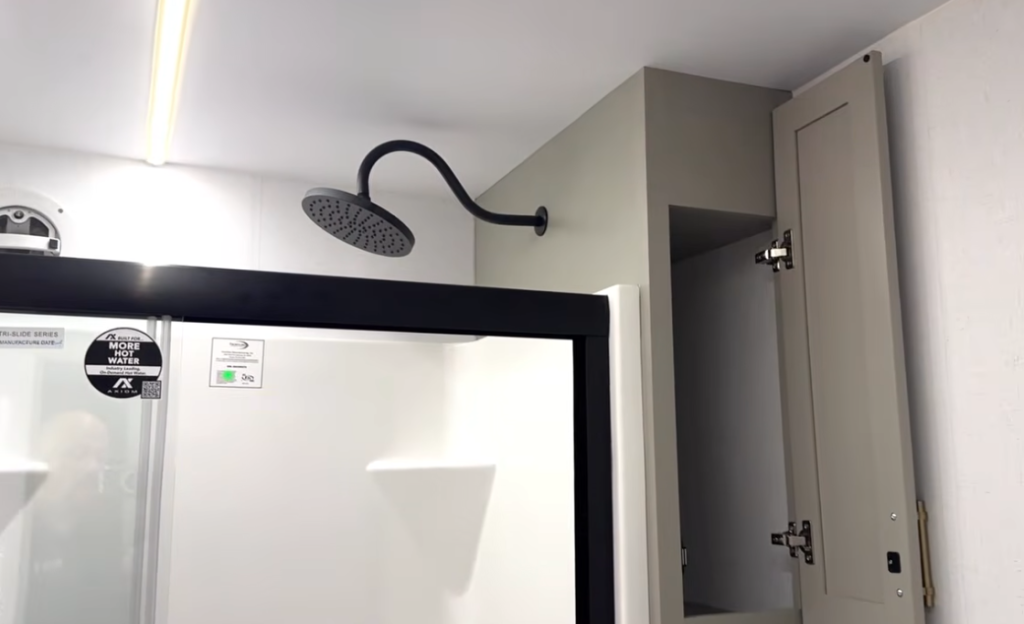 Luxury bathroom shower in the 2026 Axiom Vendetta 4250 fifth wheel featuring a black rainfall showerhead, glass sliding doors, and built-in linen storage with soft-close cabinetry.
