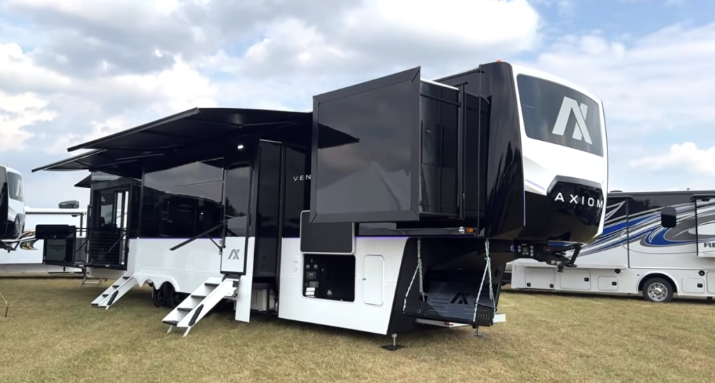 2026 Axiom Vendetta 4250 fifth wheel toy hauler with dual awnings extended, featuring a side patio, multiple slide-outs, and sleek black-and-white exterior design showcased at an RV show.