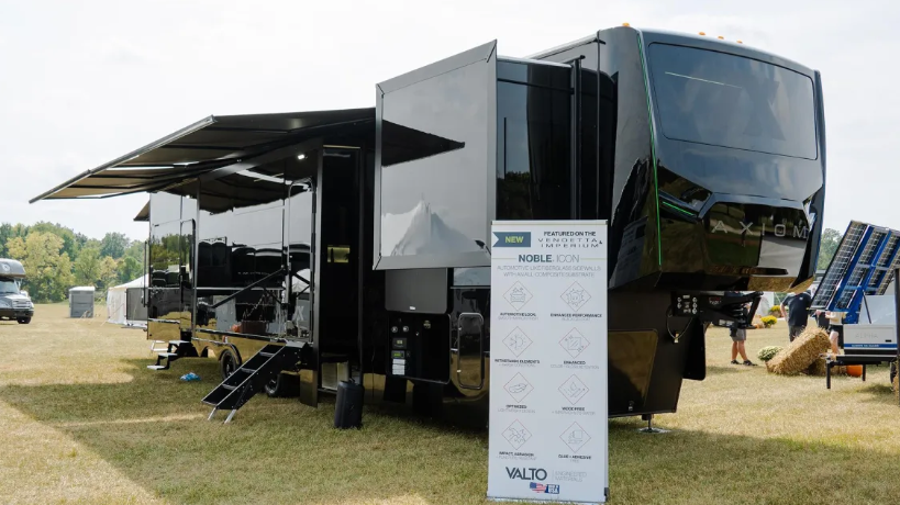 Exterior of the 2026 Axiom Vendetta 4250 luxury fifth wheel toy hauler in glossy black finish, displayed at an RV show with dual slide-outs extended, large awning, and Noble Icon automation system sign in front.