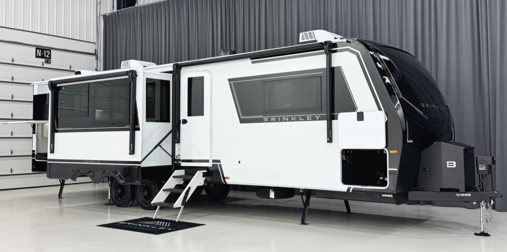 2026 Brinkley Model Z Air 315 travel trailer with triple slides extended, modern white-and-black exterior, large panoramic windows, and fold-down entry steps displayed in an indoor showroom.