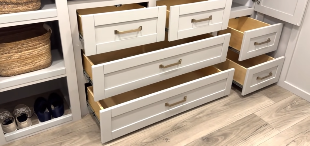 Built-in RV bedroom dresser showing multiple open soft-close drawers, white shaker cabinetry, and organized shelving with baskets and shoes on a light wood floor.